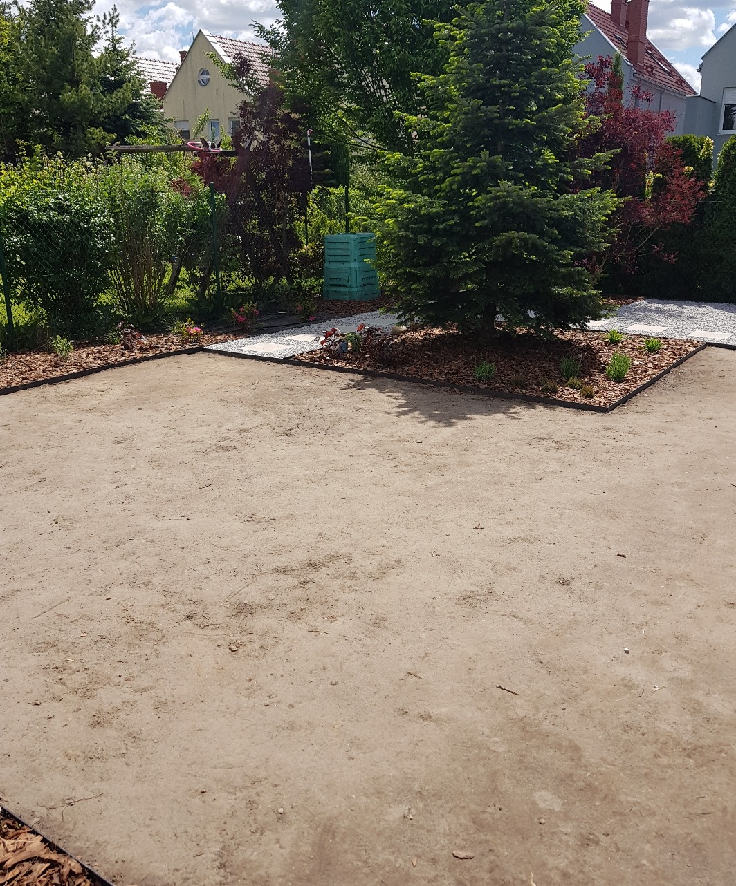 Przygotowany teren pod trawnik z obrzeżami, widoczny fragment ścieżki z kamienia i drzewo iglaste w ogrodzie.