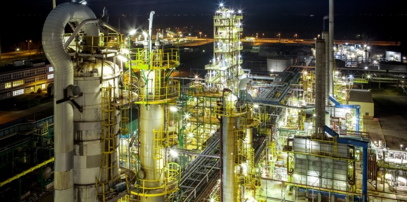 Nocny widok na rozświetloną instalację przemysłową z licznymi rurami i zbiornikami, widoczne światła uliczne i budynki w tle.