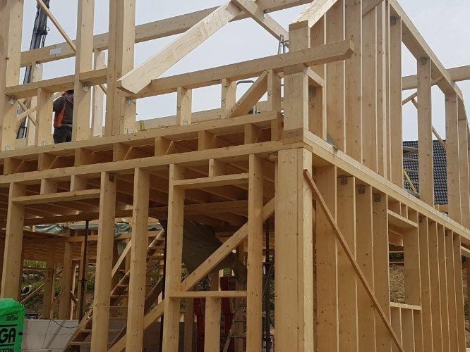 Konstrukcja szkieletowa domu w trakcie budowy, widoczne drewniane elementy nośne i pracownik na rusztowaniu w tle, zielony pakiet materiałów budowlanych na pierwszym planie.