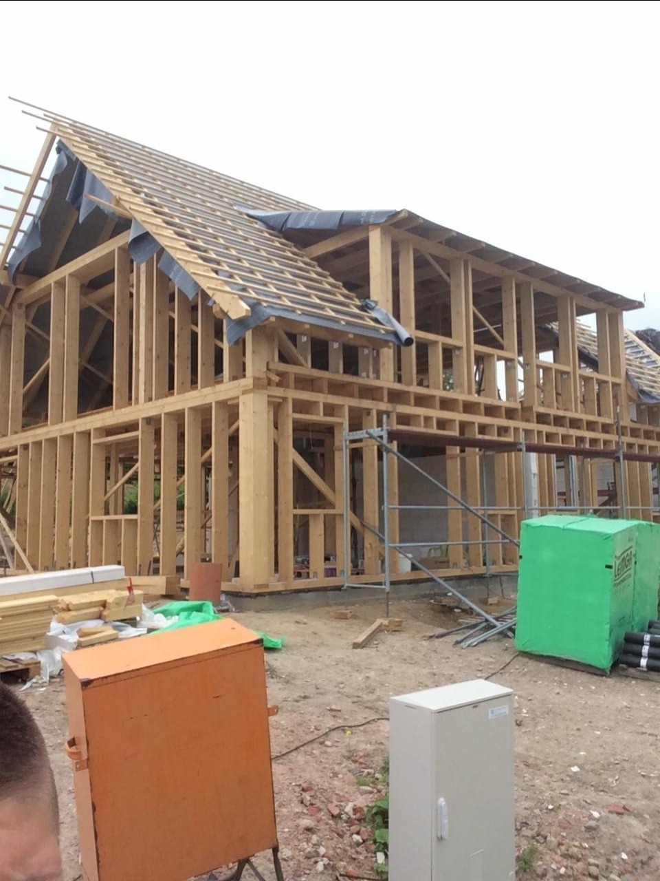 Surowa konstrukcja domu z drewna, widoczne elementy szkieletu i więźby dachowej pokryte czarną folią, rusztowanie w tle, pomarańczowa skrzynka narzędziowa i zielone paczki materiałów budowlanych...