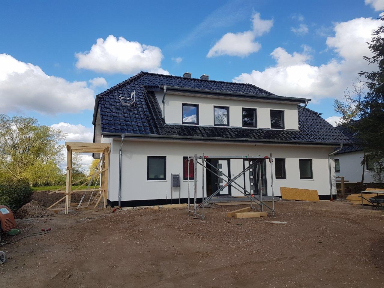 Nowo wybudowany dom jednorodzinny z ciemnym dachem, jasną elewacją i drewnianą konstrukcją dobudówki, widoczne rusztowania i teren budowy.