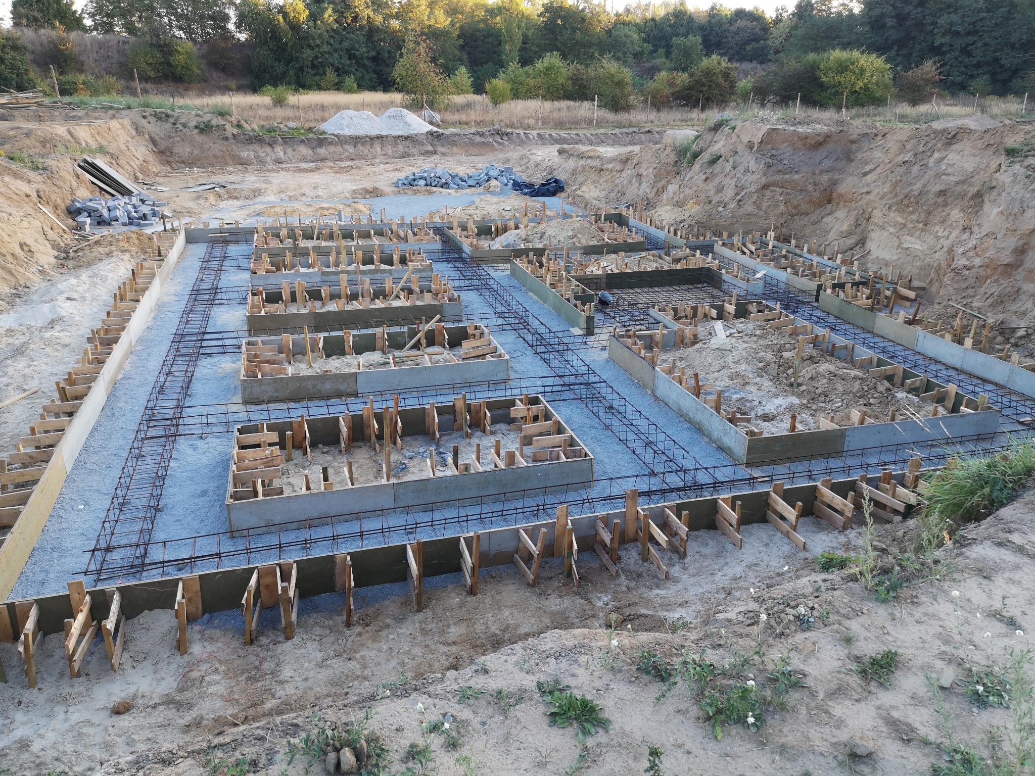 Przygotowanie zbrojenia i szalunków pod fundamenty domu jednorodzinnego na działce z wykopem, widok z góry na konstrukcję z betonu, stali i drewna.