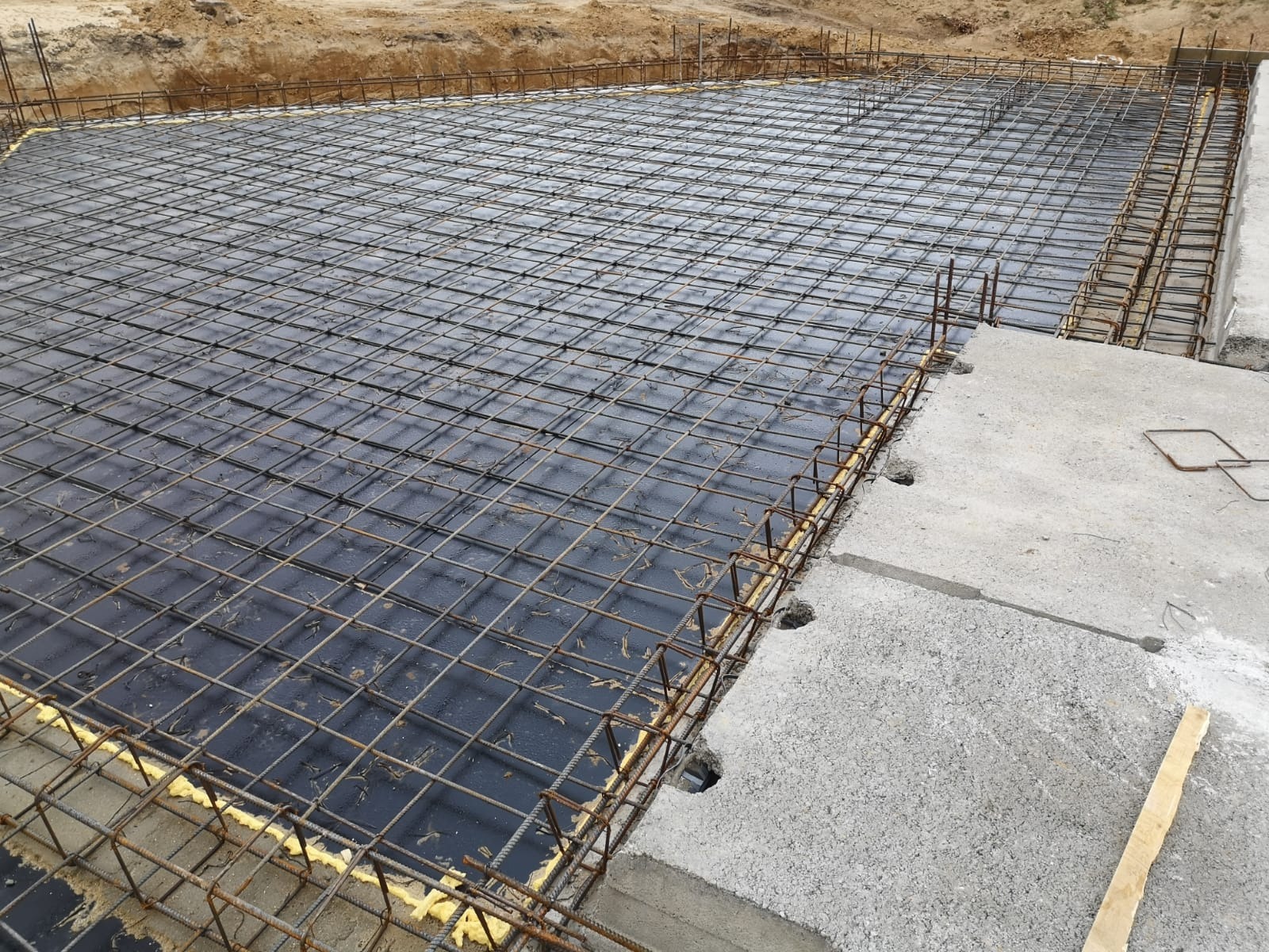 Zbrojenie płyty fundamentowej na czarnej folii, z widoczną żółtą pianką izolacyjną na obrzeżach i betonową wylewką po prawej stronie, przygotowane do zalania betonem.