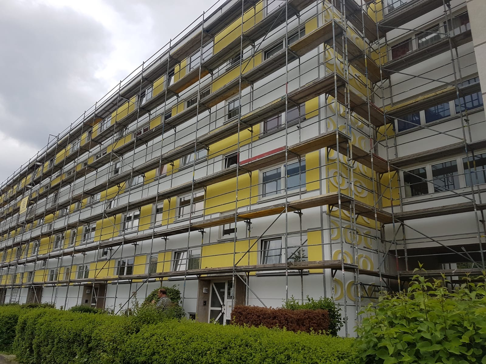 Wielopiętrowy budynek mieszkalny w trakcie prac elewacyjnych, pokryty rusztowaniem, z widocznymi fragmentami żółtej i białej elewacji oraz ozdobnymi elementami.