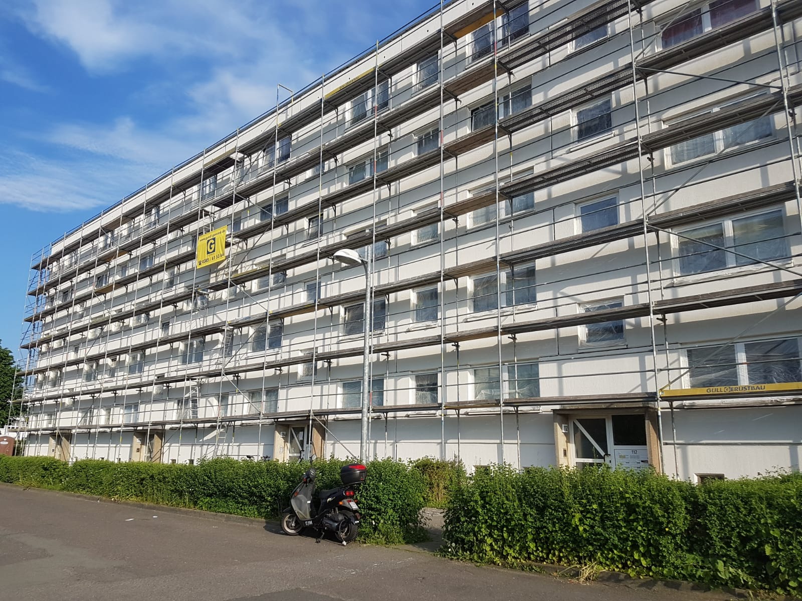 Wielopiętrowy budynek w trakcie prac elewacyjnych, osłonięte okna, rusztowania na całej fasadzie, skuter zaparkowany obok żywopłotu.