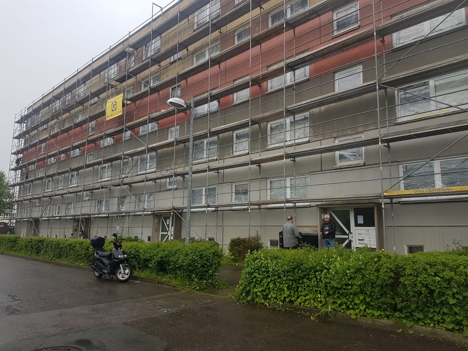 Wielopiętrowy budynek w trakcie prac elewacyjnych, osłonięte okna, rusztowania na całej fasadzie, widoczny baner reklamowy firmy na rusztowaniu, skuter zaparkowany przed budynkiem.