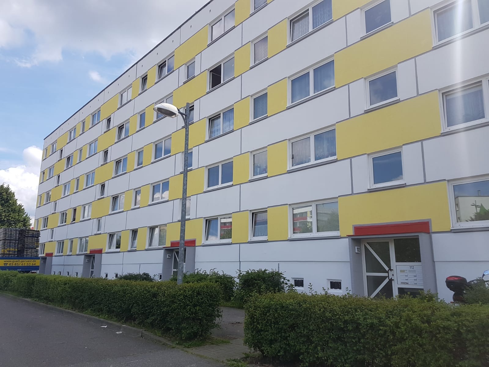 Wielopiętrowy budynek mieszkalny z elewacją w odcieniach bieli i żółci, widoczny z poziomu ulicy, z zielonym żywopłotem i słupem oświetleniowym na pierwszym planie.