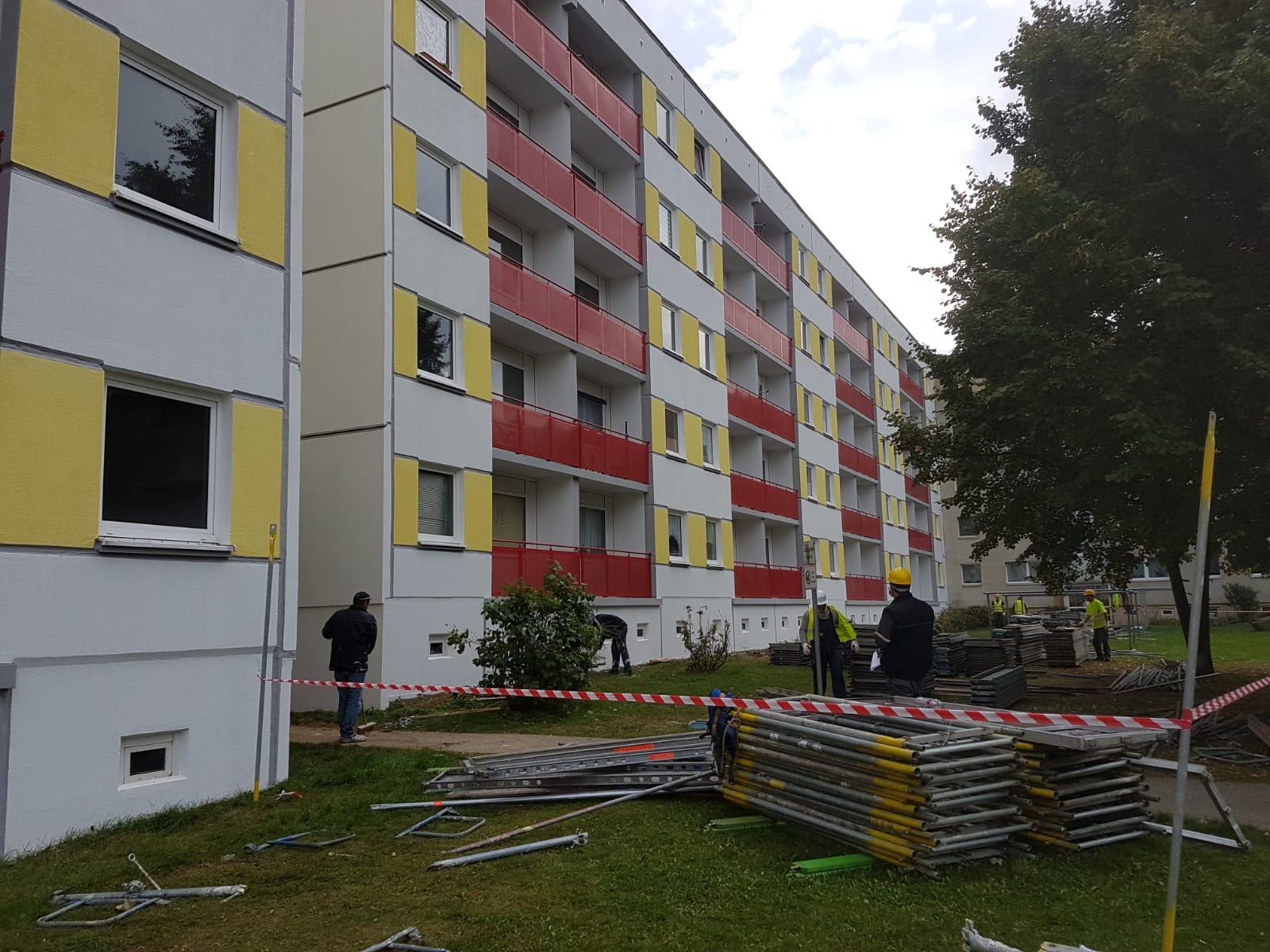 Wielopiętrowy budynek z pomalowanymi na żółto i czerwono balkonami, otoczony przez robotników w kaskach i rozstawione rusztowania, zabezpieczony taśmą ostrzegawczą na trawniku.