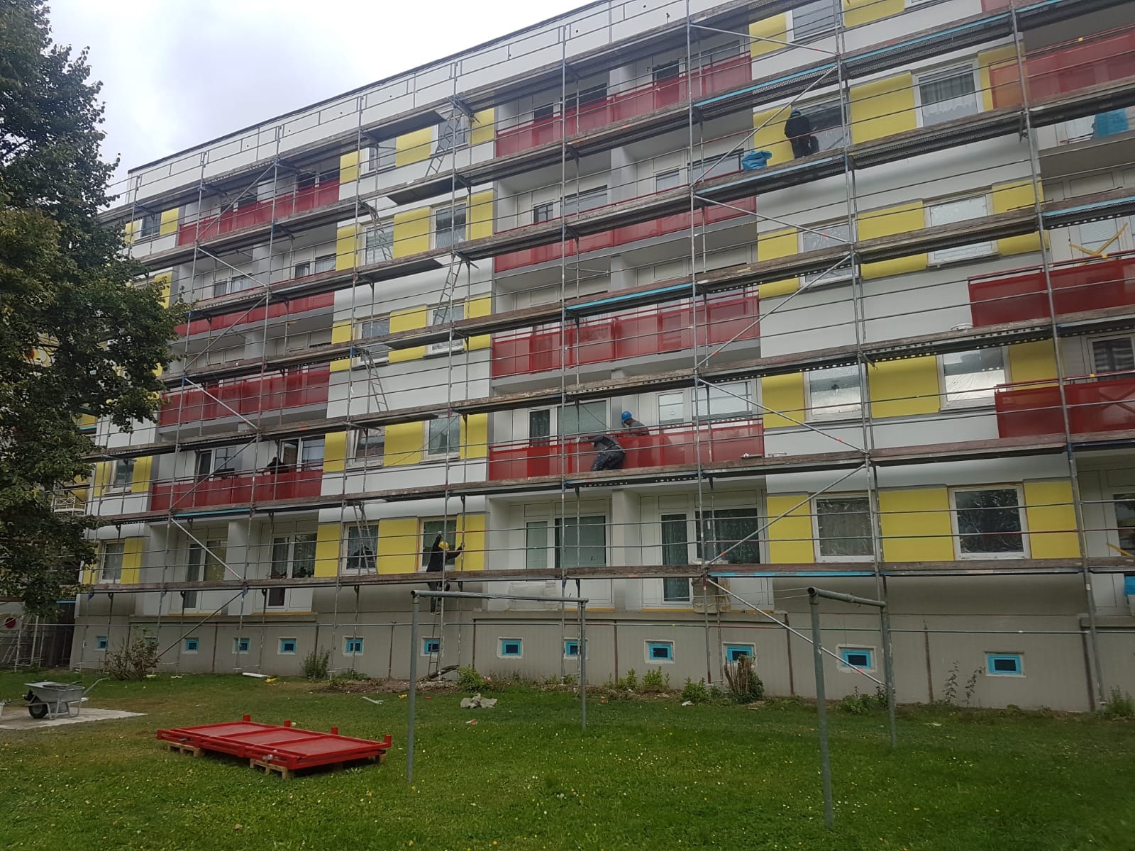 Wielopiętrowy budynek mieszkalny w trakcie prac elewacyjnych; widoczne rusztowania, robotnicy przy oknach i balkonach, pomalowane fragmenty ścian na żółto i czerwono, elementy rusztowań na trawniku.