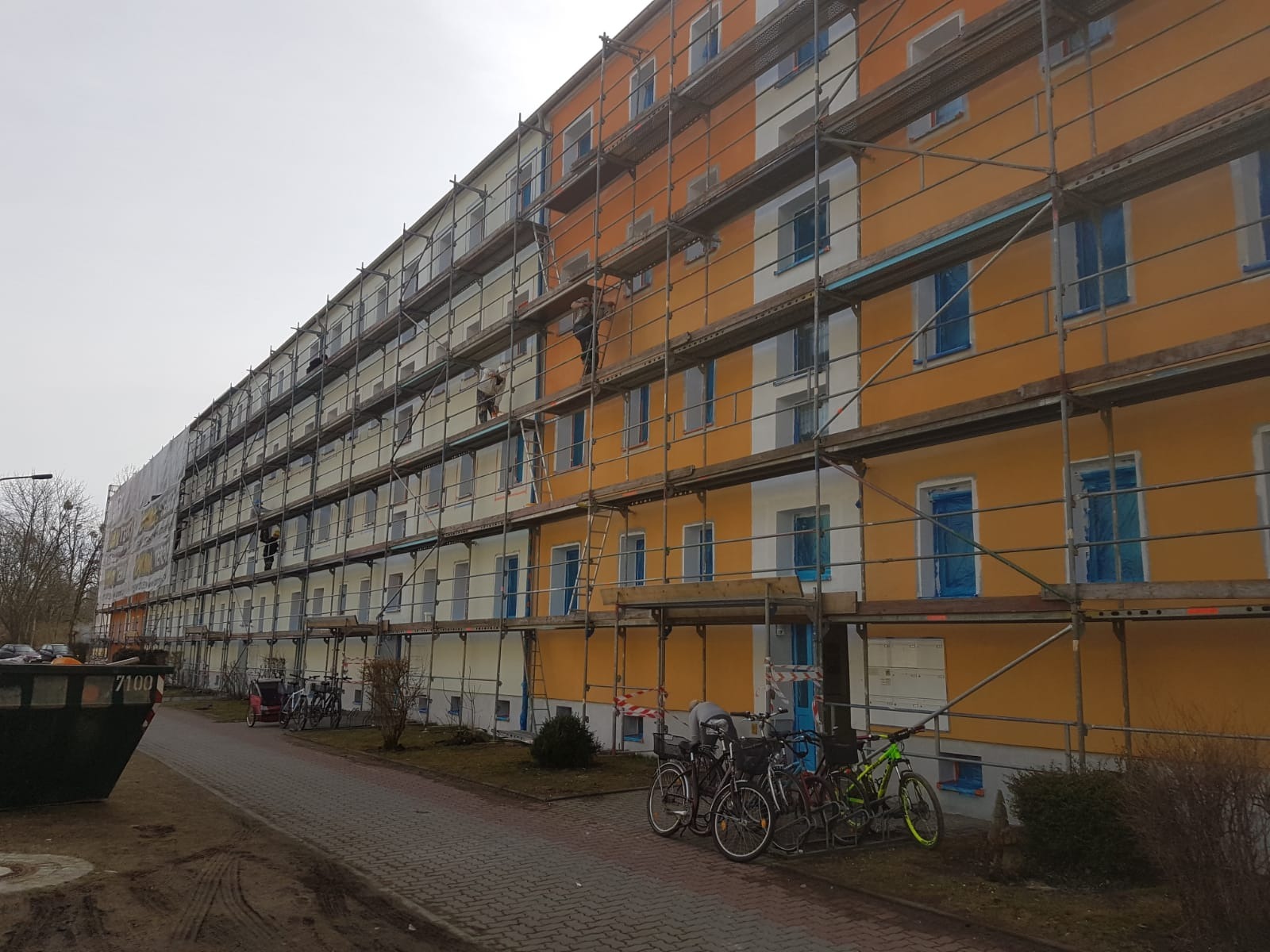 Blok mieszkalny w trakcie ocieplania elewacji, widoczne rusztowania, częściowo pomalowana fasada na pomarańczowo, okna zabezpieczone folią, na chodniku rowery i kontener na śmieci.