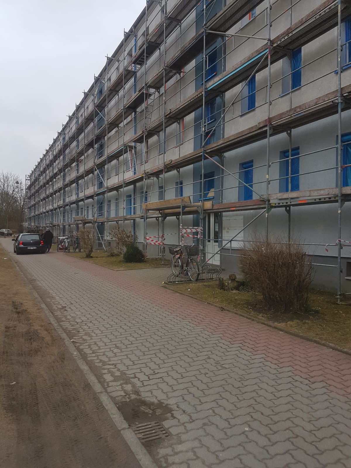 Wielopiętrowy budynek mieszkalny w trakcie prac elewacyjnych, osłonięte okna folią, rozstawione rusztowania, widoczny chodnik z kostki brukowej.