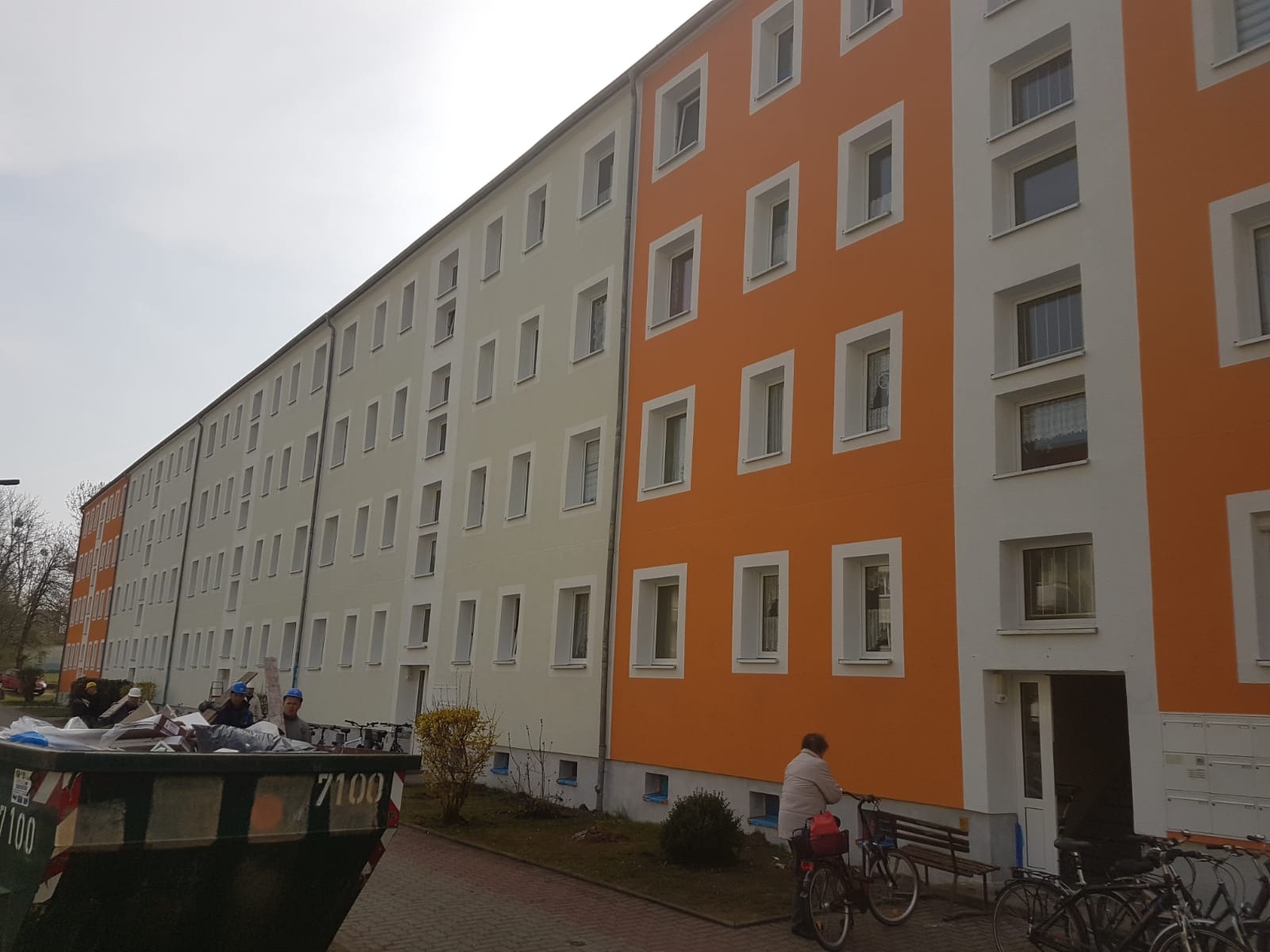 Długi blok mieszkalny po renowacji, pomalowany na pomarańczowo i jasno szaro, widoczny kontener na śmieci i pracownicy w kaskach.