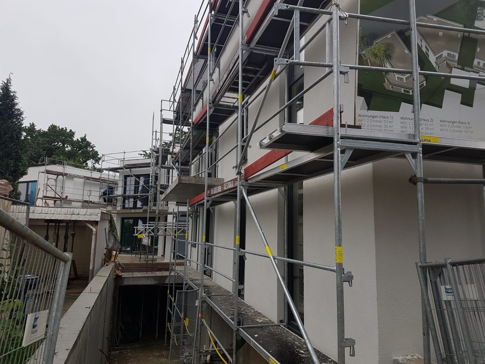 Budynek w trakcie prac wykończeniowych elewacji, z widocznym rusztowaniem i plakatem z wizualizacją projektu.