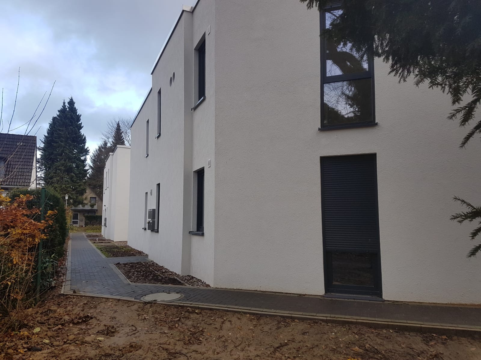 Biały budynek wielorodzinny z ciemnymi oknami i roletami, widok z zewnątrz na elewację i chodnik z kostki.