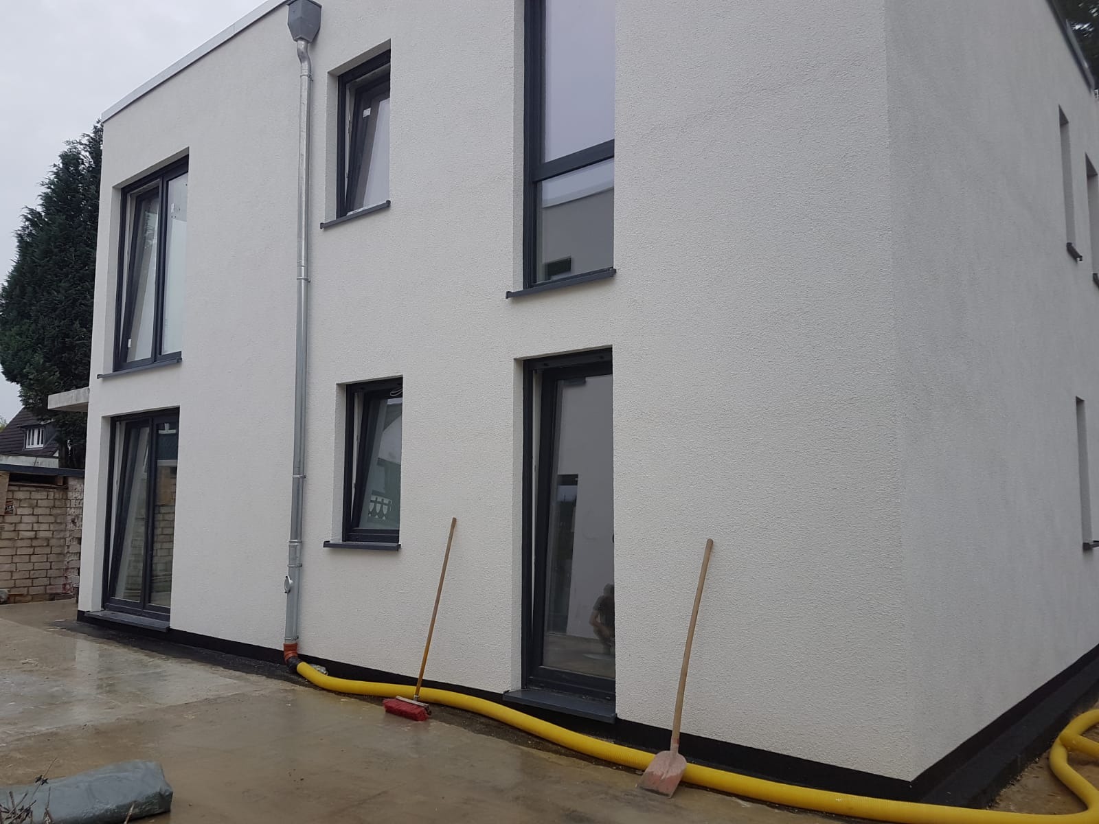 Świeżo otynkowana, biała elewacja budynku z ciemnymi oknami, widoczne rury spustowe i żółty wąż do odprowadzania wody, dwie łopaty oparte o ścianę.