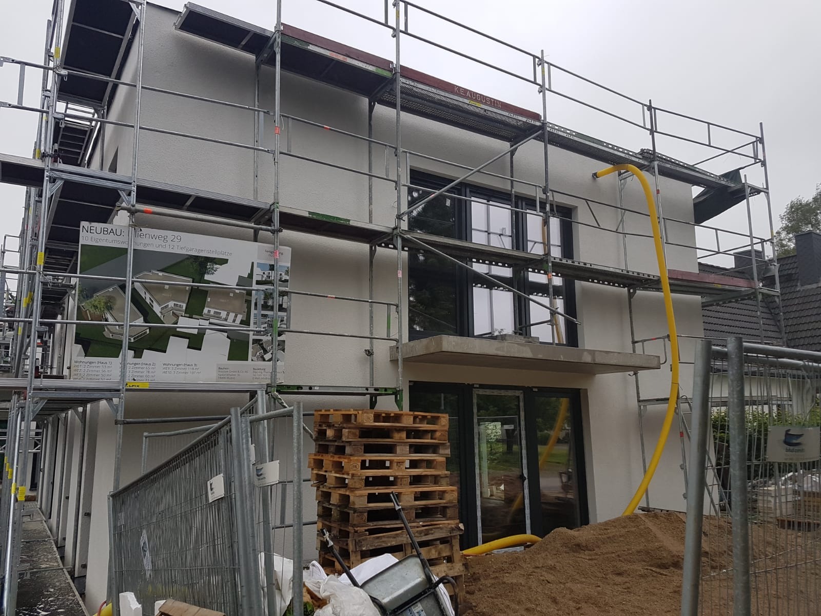 Dom w trakcie budowy z rusztowaniem, widoczny plakat informacyjny z wizualizacją budynku, stos palet i żółty wąż.