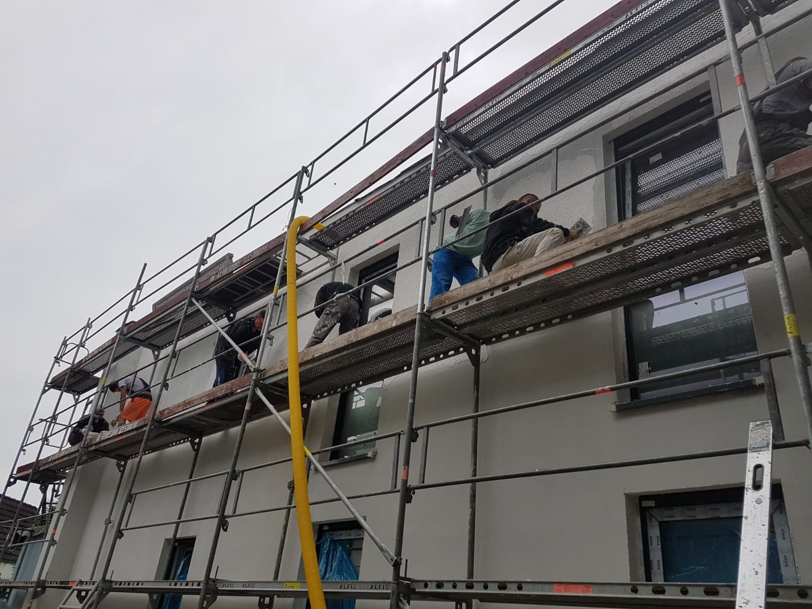 Praca na rusztowaniu: czterech robotników nakłada tynk na elewację domu jednorodzinnego, widoczny żółty wąż i okna z ciemnymi ramami.