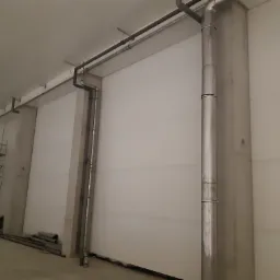 Wnętrze hali w trakcie instalacji rur wentylacyjnych, widoczne rury pionowe i poziome, drabina i elementy montażowe.