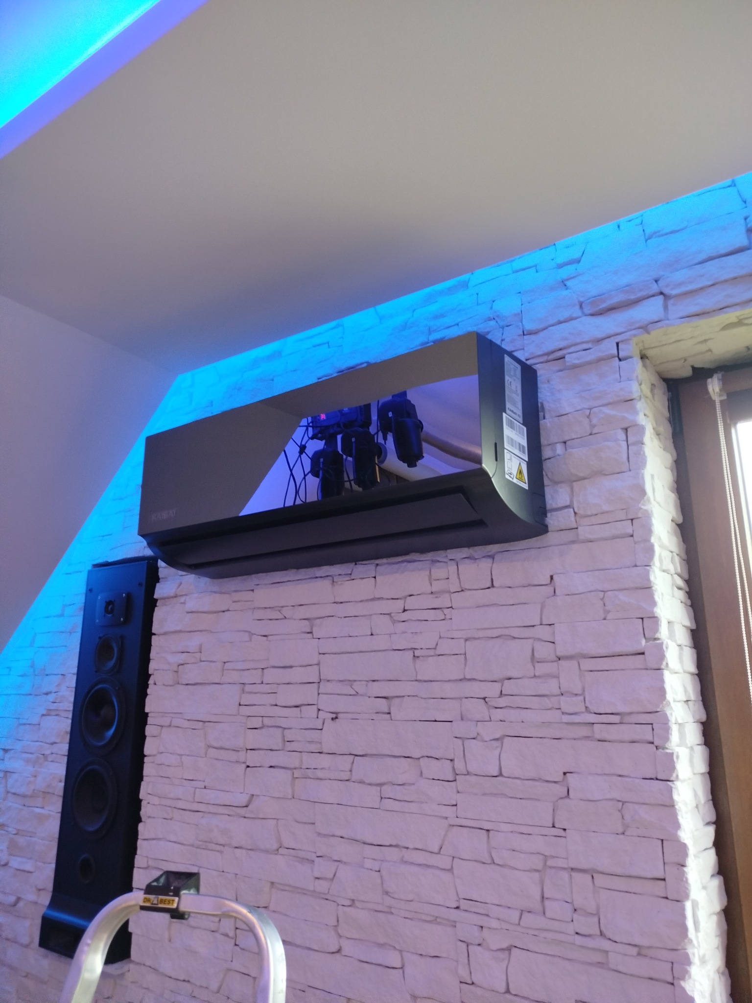 Czarny klimatyzator marki Kaisai zamontowany na ścianie z białej cegły, oświetlony niebieskim światłem LED, obok głośnika i drabiny.