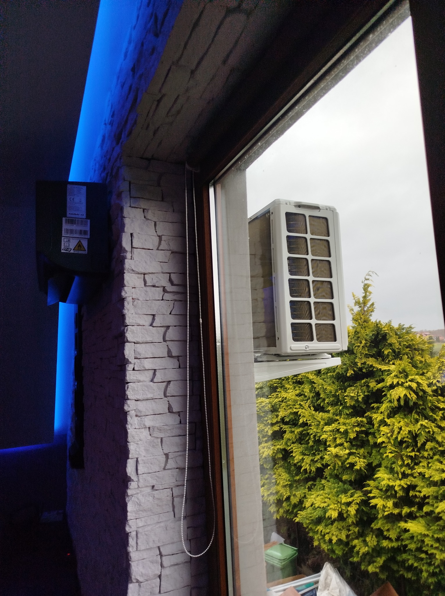 Jednostka zewnętrzna klimatyzatora zamontowana na ścianie budynku, widoczna przez okno, z zielonym krzewem w tle i ścianą z dekoracyjnej cegły wewnątrz pomieszczenia, oświetloną niebieskim światłem.