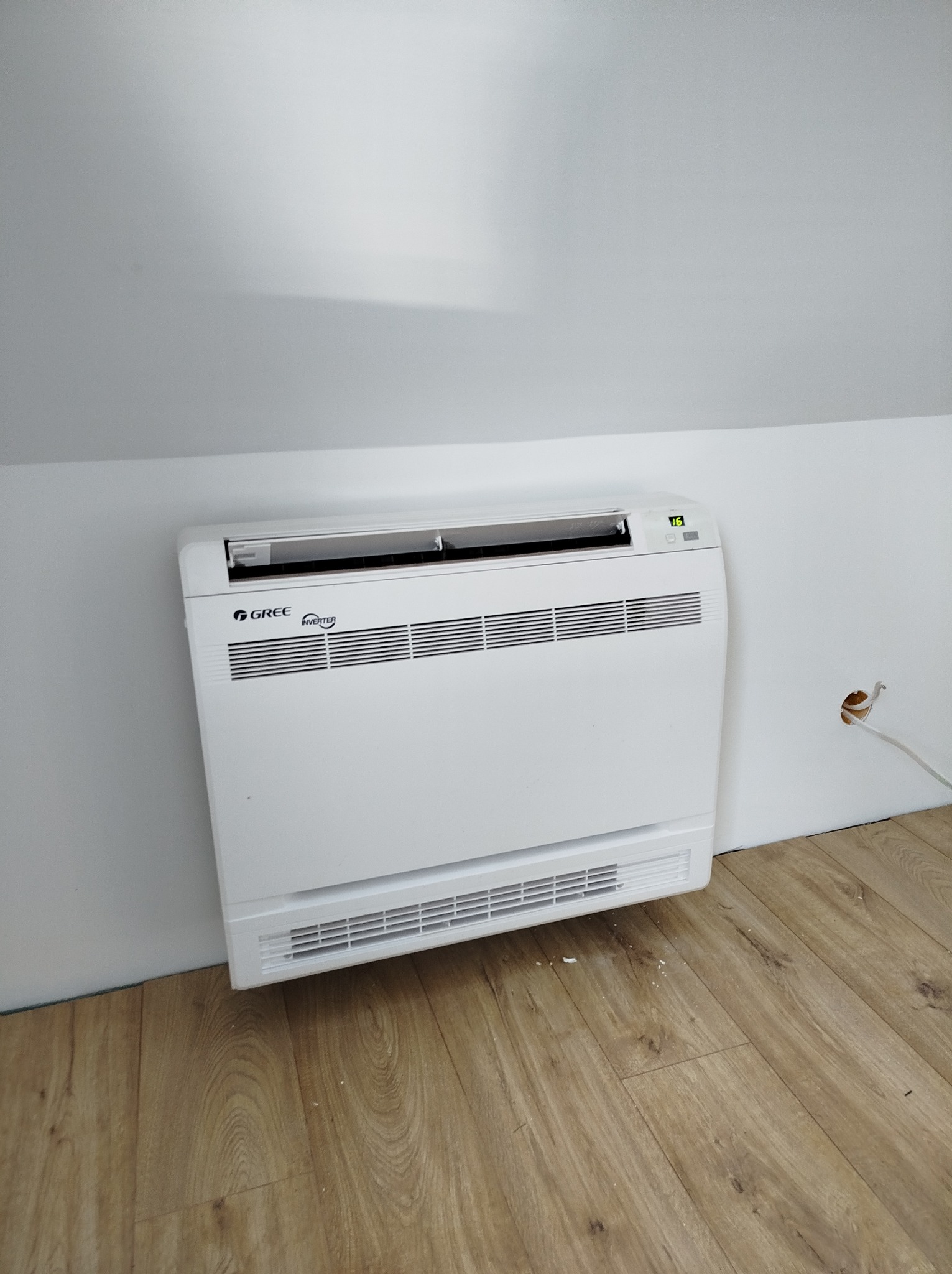 Biały klimatyzator podłogowy Gree Inverter zamontowany w pomieszczeniu ze skośnym sufitem i drewnianą podłogą, widoczny otwór w ścianie z wystającymi kablami.