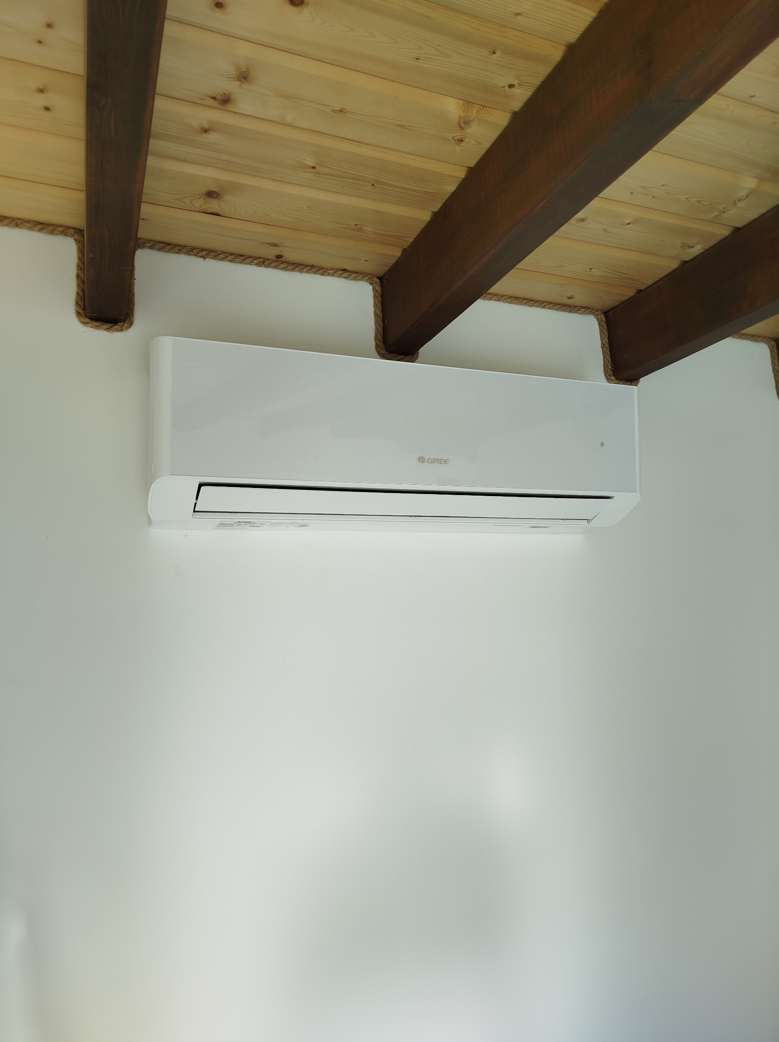 Biały klimatyzator marki Gree zamontowany na białej ścianie pod drewnianym sufitem z belkami ozdobionymi jutowym sznurem.