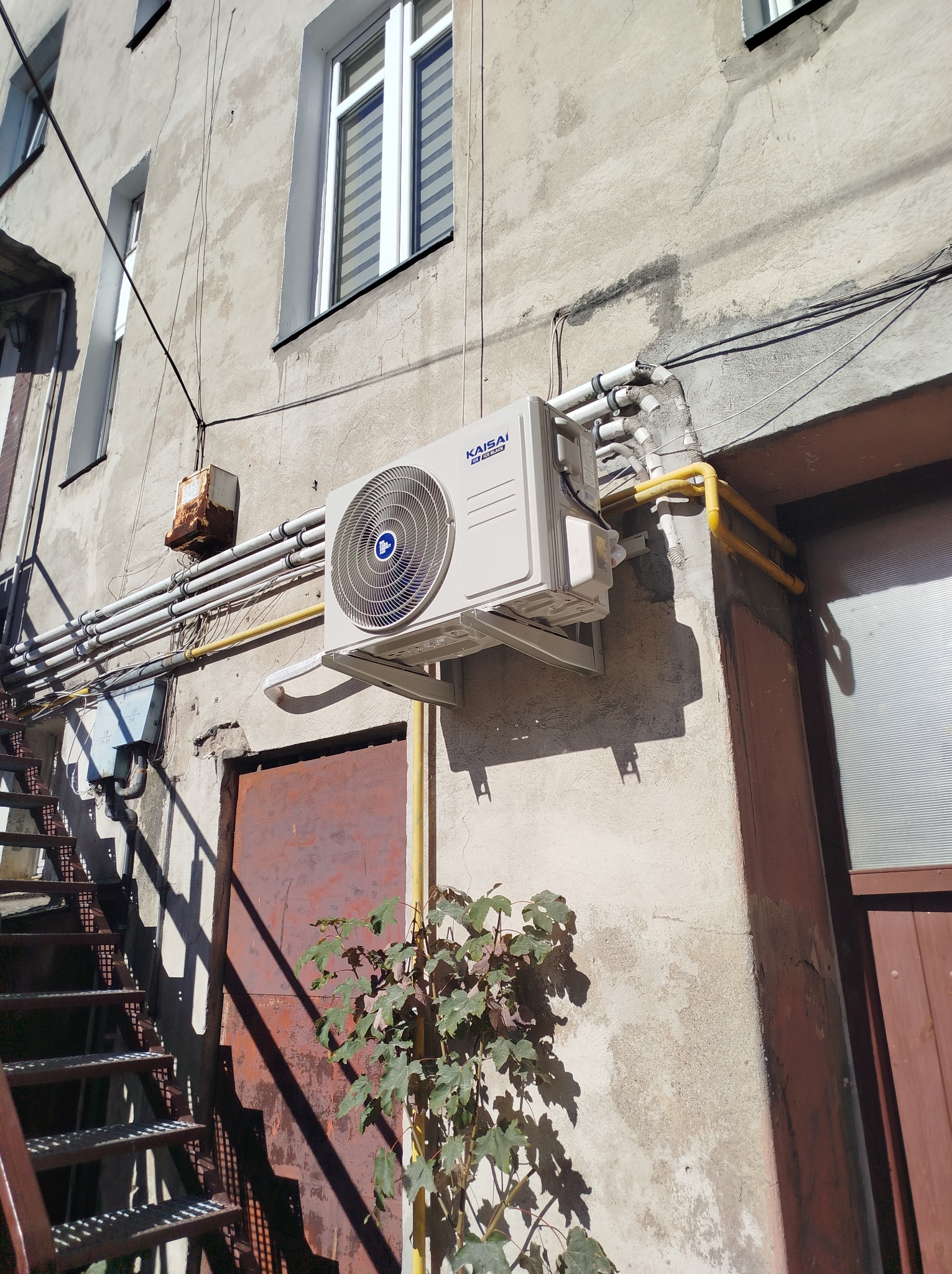 Zewnętrzna jednostka klimatyzatora Kaisai zamontowana na elewacji budynku obok metalowych schodów przeciwpożarowych i rdzewiejących rur, z młodym drzewkiem u dołu kadru.