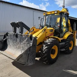 Żółta koparko-ładowarka JCB 3CX z brudną łyżką na tle szarego magazynu i błękitnego nieba z chmurami.