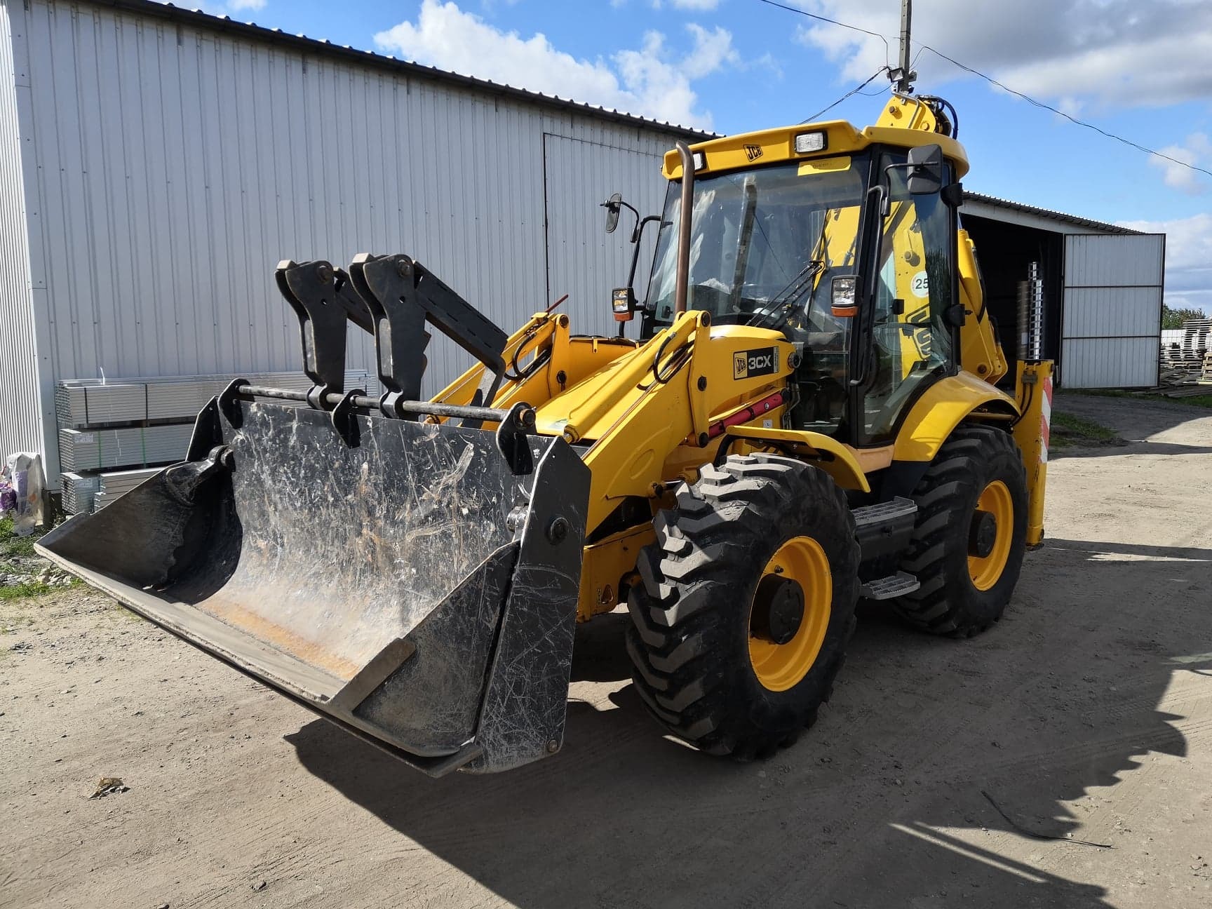 Żółta koparko-ładowarka JCB 3CX z brudną łyżką na tle szarego magazynu i błękitnego nieba z chmurami.