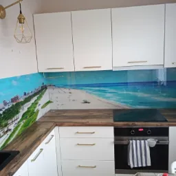 Biała kuchnia z drewnianym blatem i panelem ściennym z motywem plaży i oceanu, oświetlona złotą lampą wiszącą.