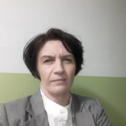 Portret kobiety w szarym żakiecie i białej koszuli na tle ściany w dwóch odcieniach zieleni.