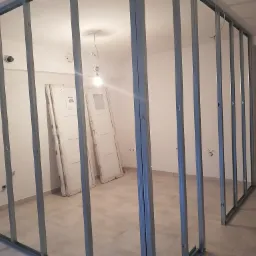 Metalowa konstrukcja szkieletowa ścian działowych w trakcie montażu w jasnym pomieszczeniu; widoczne przygotowane do montażu białe drzwi i surowa instalacja elektryczna.