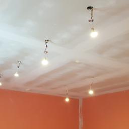 Usługi remontowo - budowlane NerDzik - Niedokończony sufit z płyt gipsowo-kartonowych z widocznymi łączeniami i prowizorycznym oświetleniem w pomieszczeniu z pomarańczowymi ścianami. Widoczne kable i żarówki wiszące z otworów w suficie.