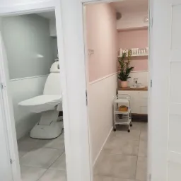 Widok przez otwarte drzwi na gabinet kosmetyczny z białym fotelem zabiegowym, w tle ściana w kolorze pudrowego różu, biała szafka i wózek kosmetyczny.