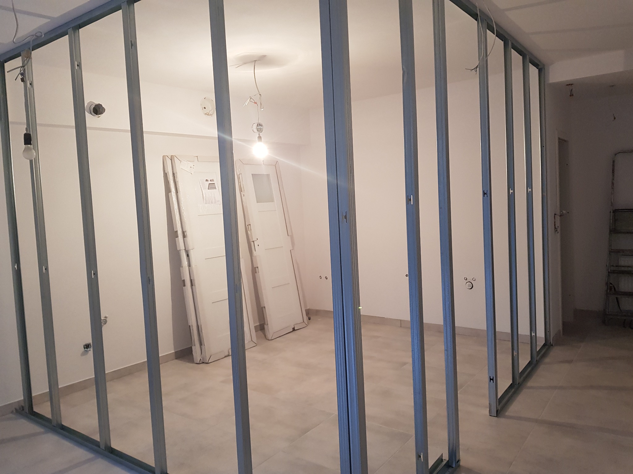 Metalowa konstrukcja szkieletowa ścian działowych w trakcie montażu w jasnym pomieszczeniu; widoczne przygotowane do montażu białe drzwi i surowa instalacja elektryczna.