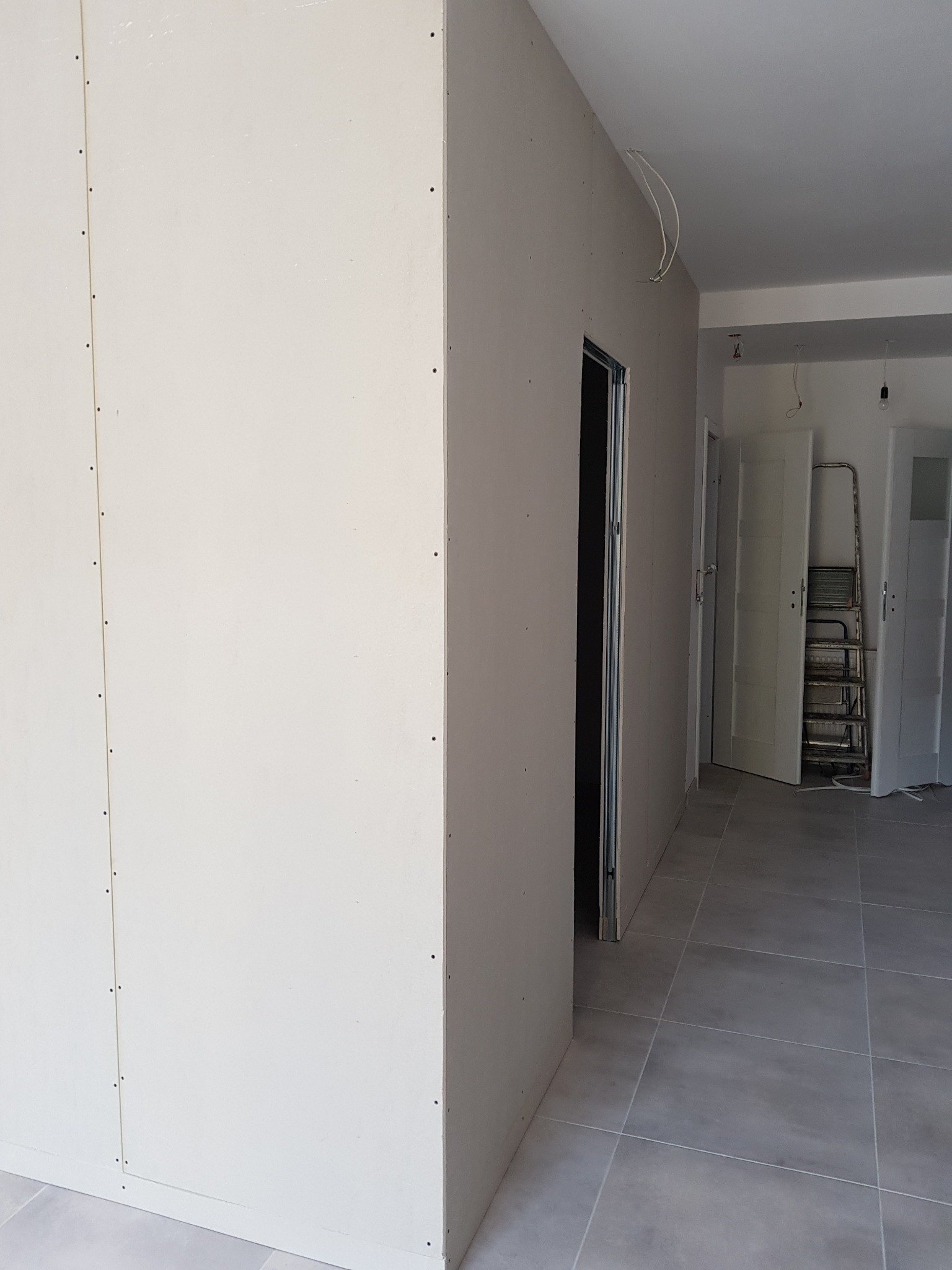 Wnętrze w trakcie remontu: ściany wykończone płytami gipsowo-kartonowymi, widoczne łączenia i wkręty, szara podłoga z płytek, futryna drzwiowa, w tle drabina oparta o ścianę i wiszące przewody...