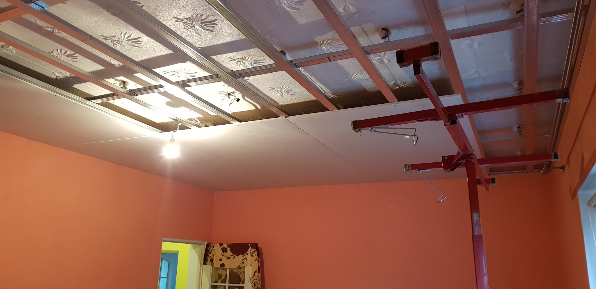 Montaż sufitu podwieszanego z widoczną konstrukcją metalową i podnośnikiem w pomieszczeniu z pomarańczowymi ścianami, oświetlonym żarówką.