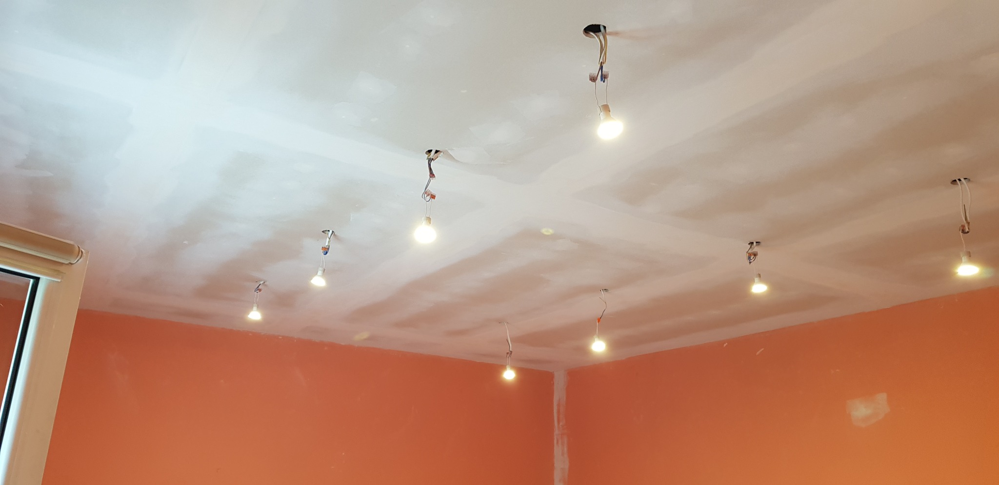 Niedokończony sufit z płyt gipsowo-kartonowych z widocznymi łączeniami i prowizorycznym oświetleniem w pomieszczeniu z pomarańczowymi ścianami. Widoczne kable i żarówki wiszące z otworów w suficie.