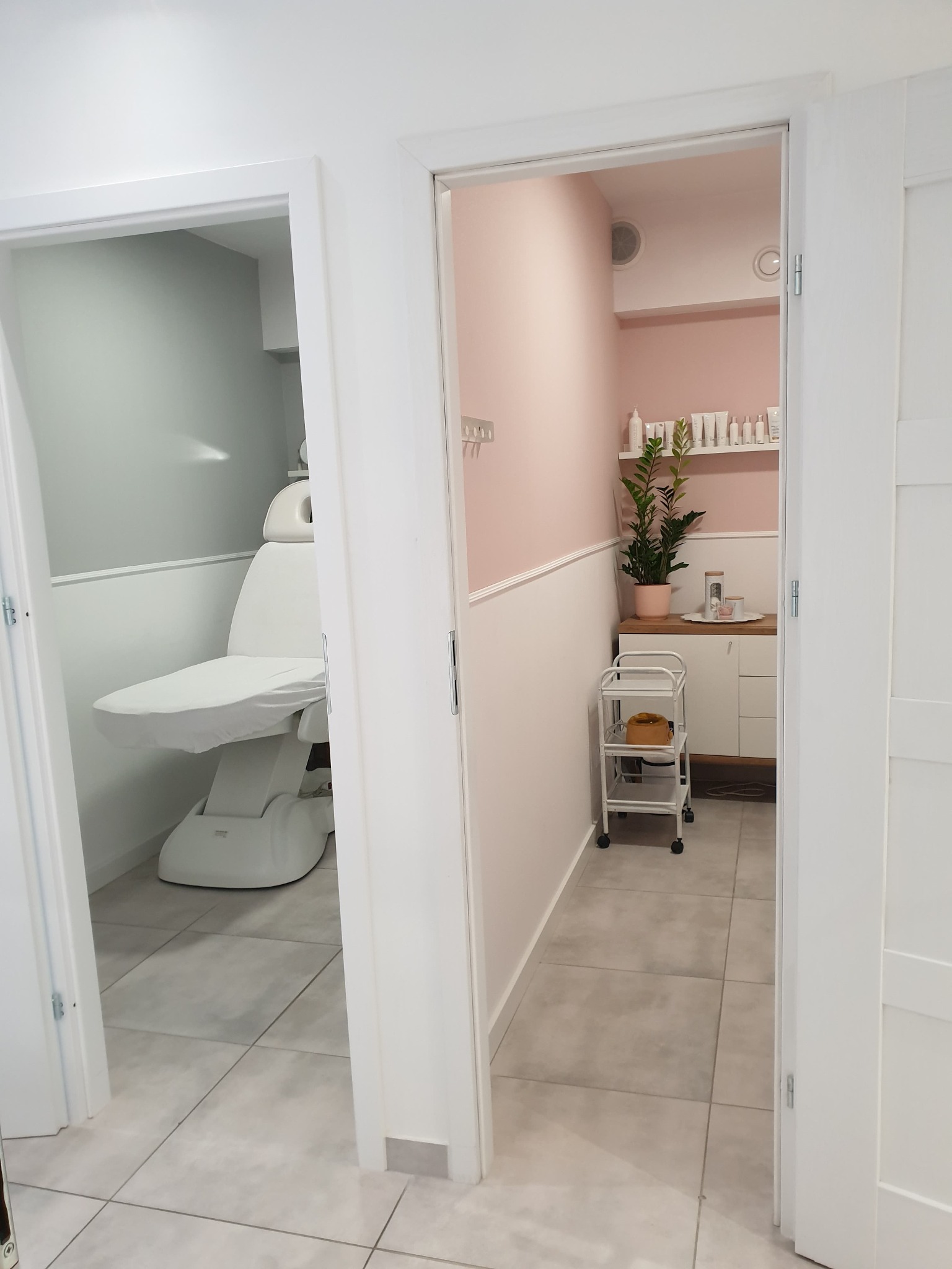 Widok przez otwarte drzwi na gabinet kosmetyczny z białym fotelem zabiegowym, w tle ściana w kolorze pudrowego różu, biała szafka i wózek kosmetyczny.
