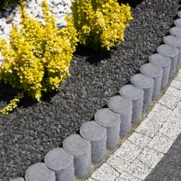 Obrzeże z betonowych palisad oddzielające nawierzchnię z kostki brukowej od rabaty z czarnym grysem, żółtymi krzewami i białymi kamieniami.