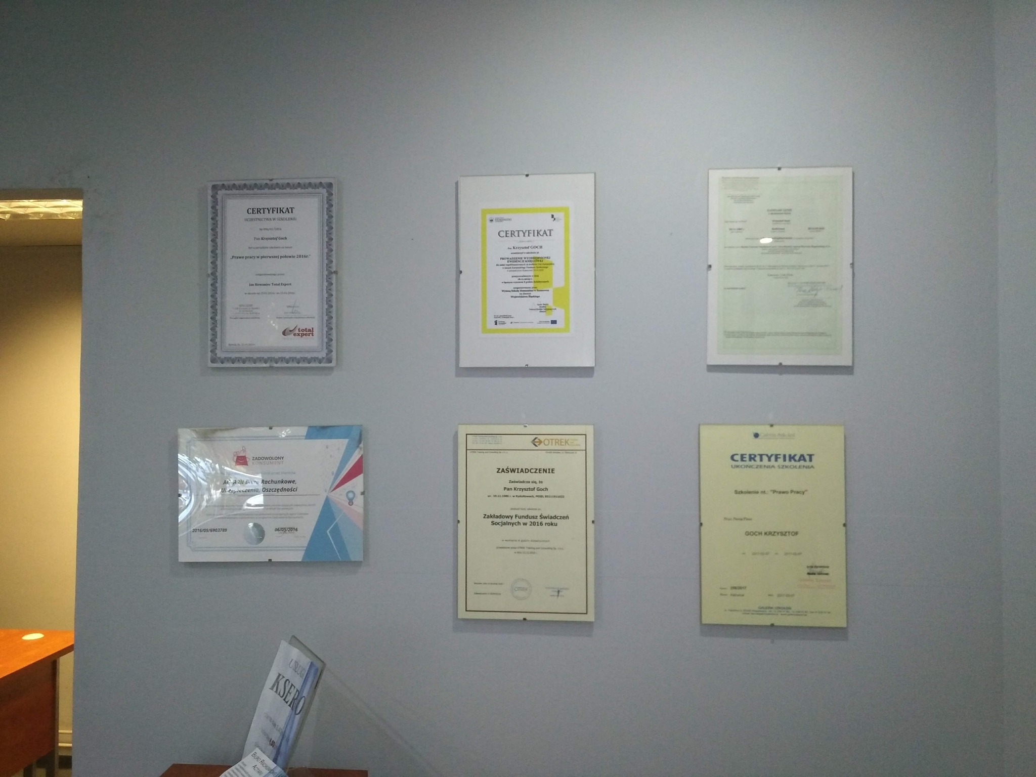 Ściana biura z powieszonymi certyfikatami i zaświadczeniami, w tym certyfikaty ukończenia szkoleń i zaświadczenia o udziale w programach socjalnych, obok stoi ulotka reklamowa z napisem 'Usługi...