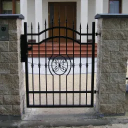 Czarna, metalowa furtka z dekoracyjnym wzorem, osadzona w kamiennych słupkach, prowadząca do domu z brązowymi drzwiami wejściowymi i schodami.