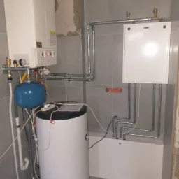 Nowoczesna instalacja grzewcza z białym kotłem, zbiornikiem ciepłej wody i rozdzielaczem, rury poprowadzone wzdłuż szarej ściany z płytek, widoczne elementy montażowe.