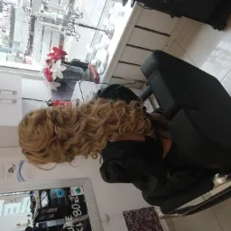 Klientka z długimi, blond, kręconymi włosami siedzi w fotelu fryzjerskim w salonie, widok zza pleców, w tle okno z dekoracjami.