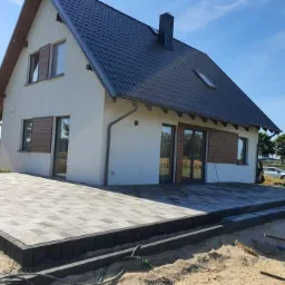 Nowoczesny dom z tarasem z szarej kostki brukowej obramowanym czarnym obrzeżem, widok z poziomu gruntu na częściowo zagospodarowany teren.