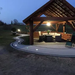 Oświetlona ścieżka z kostki brukowej prowadząca do drewnianej altany z meblami ogrodowymi, widok wczesnym wieczorem.