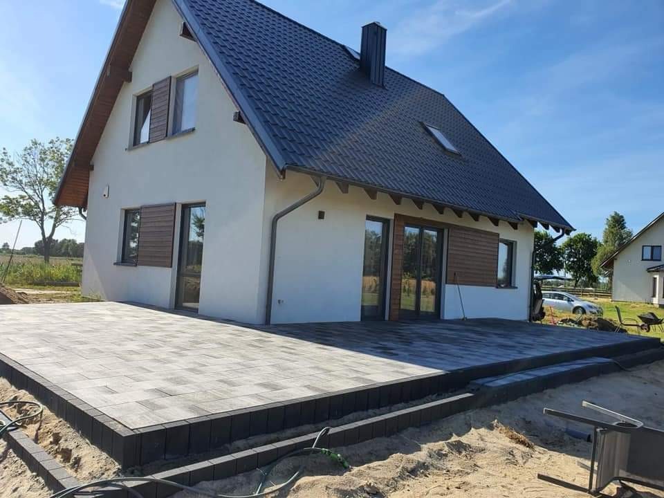 Nowoczesny dom z tarasem z szarej kostki brukowej obramowanym czarnym obrzeżem, widok z poziomu gruntu na częściowo zagospodarowany teren.