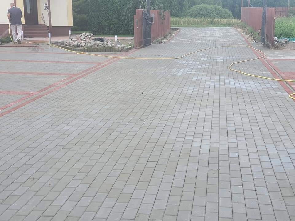 Szeroki podjazd wyłożony szarą kostką brukową z czerwonymi obramowaniami, widoczny fragment domu, brama wjazdowa i osoba z wężem ogrodowym.