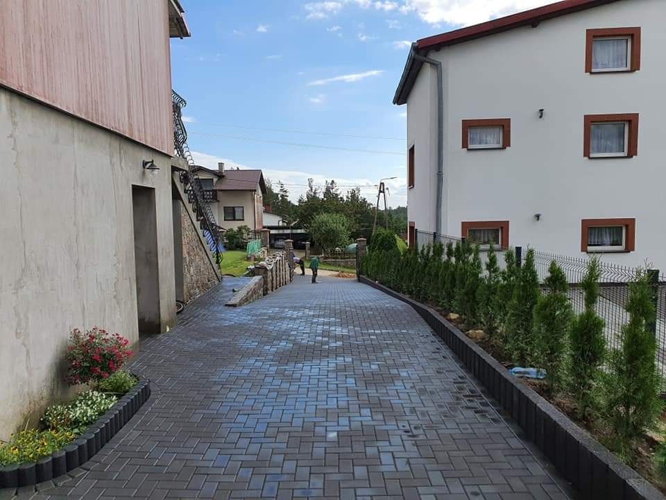 Ułożona szara kostka brukowa prowadząca do domu, z obrzeżami, młodymi iglakami i kwiatami, dwóch pracowników w oddali.