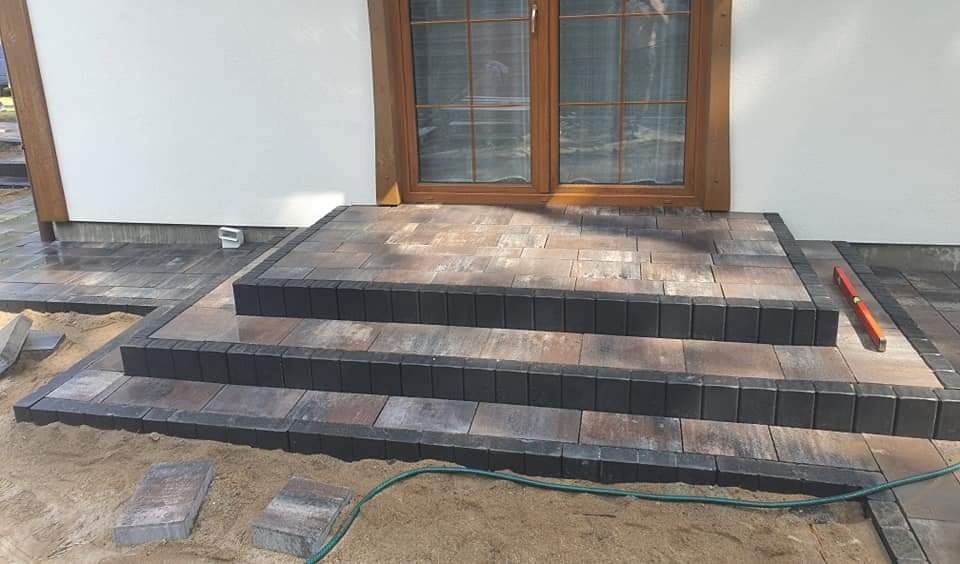 Trzy stopnie schodów wejściowych wyłożone kostką brukową w odcieniach brązu i szarości, obrzeża z ciemnej kostki, poziomica leżąca obok, widoczne piasek i wąż ogrodowy.